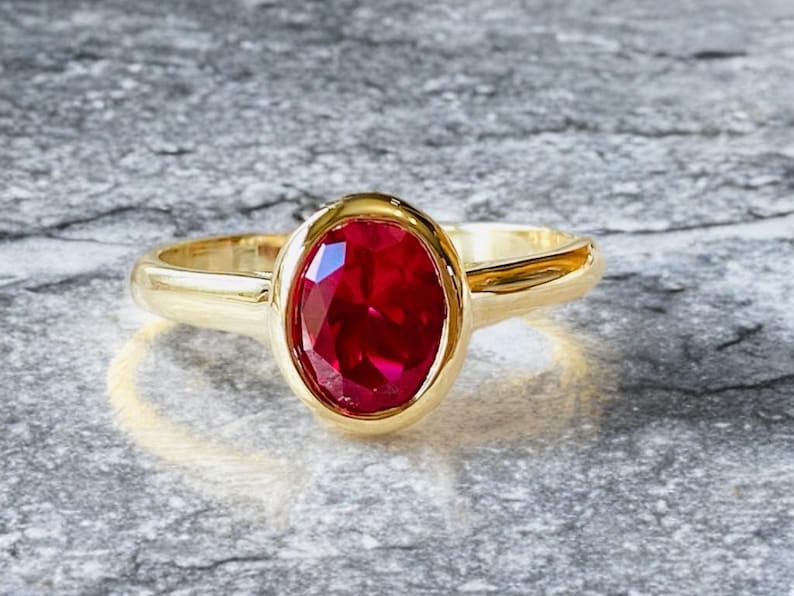 K&ouml;nnte beinhalten: Ein goldener Ring mit einem gro&szlig;en, oval geschliffenen Rubin. Der Rubin hat eine leuchtend rote Farbe und ist in einem polierten Goldband gefasst. Der Ring wird auf einer strukturierten grauen Oberfl&auml;che gezeigt.