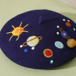 May include: A navy blue beret with a felt texture, embroidered with a yellow sun, planets, and stars. The planets are embroidered in different colors, including blue, yellow, and orange.