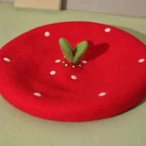 May include: A red beret hat shaped like a strawberry. The hat features white seed-like dots and green felt leaves with small green beads. The beret is a playful accessory, perfect for themed outfits or adding a touch of whimsy to any look.