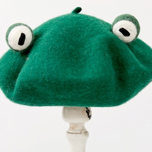 Puede incluir: Un gorro de fieltro verde con dos grandes ojos de fieltro blanco y pupilas negras. El gorro está sobre un soporte blanco.