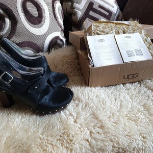 Puede incluir: Un par de sandalias de tacón de cuero negro con puntera abierta y una hebilla. Las sandalias están sobre una alfombra blanca y esponjosa. Las sandalias están en una caja con el logotipo de UGG Australia.