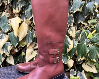 May include: A pair of tall, reddish-brown leather boots. The boots have a knee-high design with a flat sole and two decorative buckles on the side. The boots are set against a backdrop of green and yellow foliage.