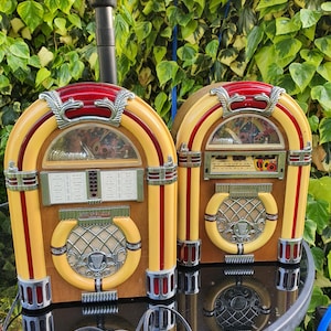 May include: Two retro jukeboxes with a yellow and red arched top, cream-coloured front, and wooden base. The jukeboxes are on a reflective surface, with a green leafy background. The design is reminiscent of the 1950s.