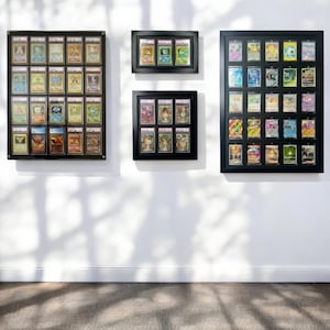 May include: Three black framed displays of collectible trading cards mounted on a white wall. The displays vary in size and contain multiple cards, each encased in a protective holder. The cards feature colourful illustrations and text.