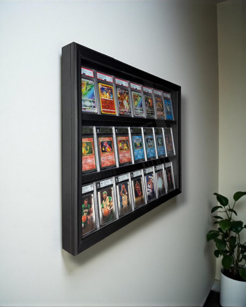 K&ouml;nnte beinhalten: Ein schwarzer, gerahmter Display-Kasten an einer wei&szlig;en Wand, der Reihen von Sammelkarten zeigt. Die Karten sind in Schutzh&uuml;llen untergebracht und zeigen verschiedene farbenfrohe Designs und Bilder. Der rechteckige Kasten hat mehrere Regale.