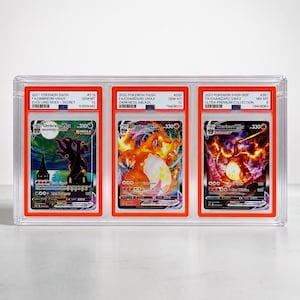 May include: A clear plastic display case holding three graded Pok&eacute;mon cards. The cards feature Umbreon VMAX and two Charizard VMAX cards. Each card is encased in a protective holder with grading information visible.