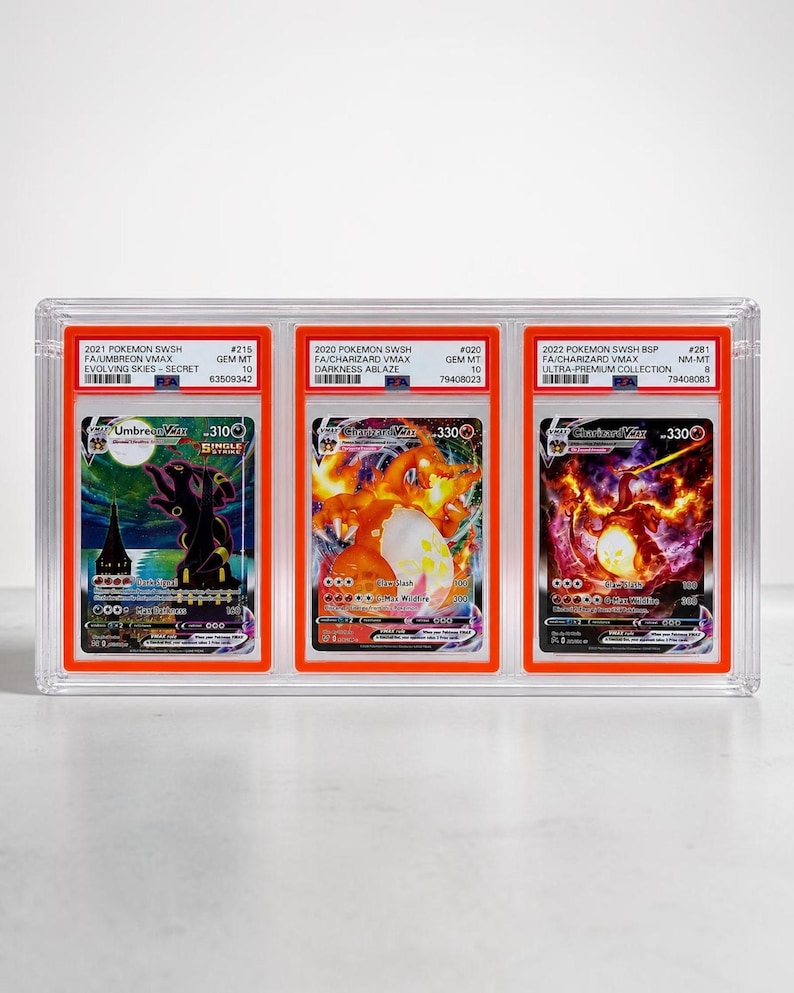 May include: A clear plastic display case holding three graded Pok&eacute;mon cards. The cards feature Umbreon VMAX and two Charizard VMAX cards. Each card is encased in a protective holder with grading information visible.