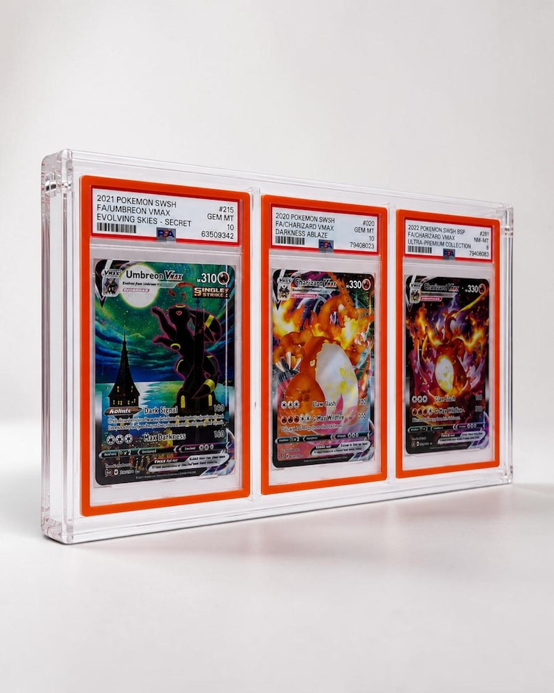 May include: A clear acrylic display case holding three graded Pok&eacute;mon trading cards. The cards feature Umbreon VMAX, Charizard VMAX, and Charizard VMAX. Each card is encased in a protective holder with grading information visible.