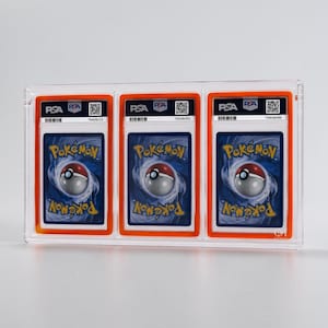 May include: A clear acrylic display case holding three graded Pok&eacute;mon trading cards. Each card is encased in a PSA holder with an orange border and features the Pok&eacute;mon logo and a barcode. The cards have a blue background with a swirling design.