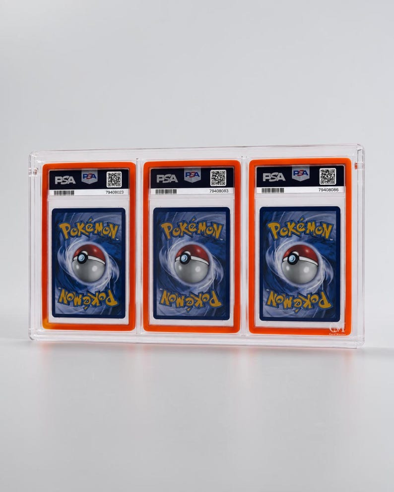 May include: A clear acrylic display case holding three graded Pok&eacute;mon trading cards. Each card is encased in a PSA holder with an orange border and features the Pok&eacute;mon logo and a barcode. The cards have a blue background with a swirling design.