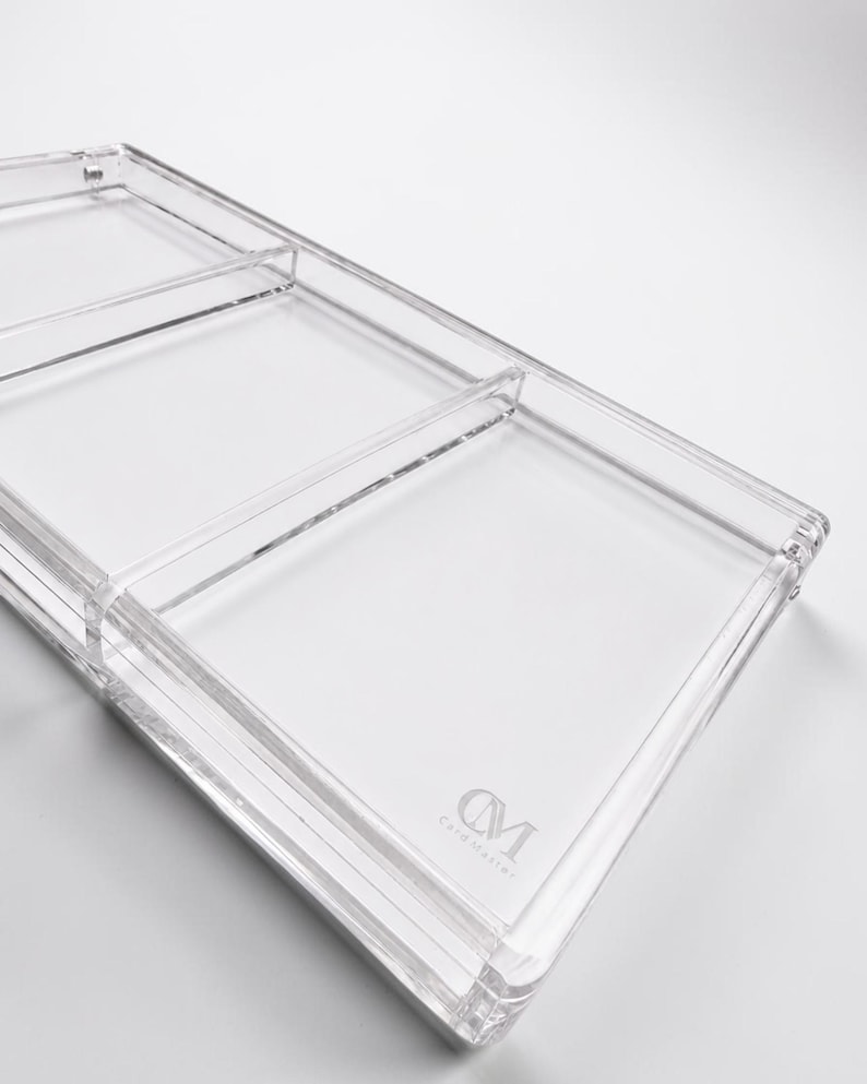 May include: A clear, rectangular acrylic tray with three compartments. The tray has rounded corners and a raised edge. The words "CM Card Master" are printed in a light gray color on the bottom right corner.