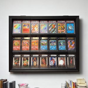 K&ouml;nnte beinhalten: Eine schwarze Vitrine an der Wand, die bewertete Sammelkarten zeigt. Die Vitrine enth&auml;lt Reihen von Pok&eacute;mon-Karten und Basketballkarten, jeweils in Schutzh&uuml;llen. Die Karten weisen leuchtende Farben und detaillierte Kunstwerke auf.