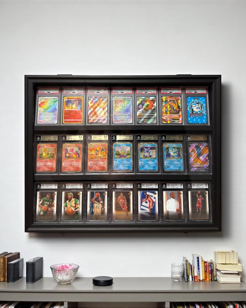 K&ouml;nnte beinhalten: Eine schwarze Vitrine an der Wand, die bewertete Sammelkarten zeigt. Die Vitrine enth&auml;lt Reihen von Pok&eacute;mon-Karten und Basketballkarten, jeweils in Schutzh&uuml;llen. Die Karten weisen leuchtende Farben und detaillierte Kunstwerke auf.