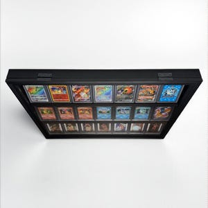 K&ouml;nnte beinhalten: Eine schwarze Vitrine mit Glasfront, die eine Sammlung farbenfroher Sammelkarten pr&auml;sentiert. Die Karten sind in Reihen angeordnet, wobei verschiedene Designs und Illustrationen sichtbar sind. Das Geh&auml;use ist rechteckig und scheint an einer wei&szlig;en Wand montiert zu sein.