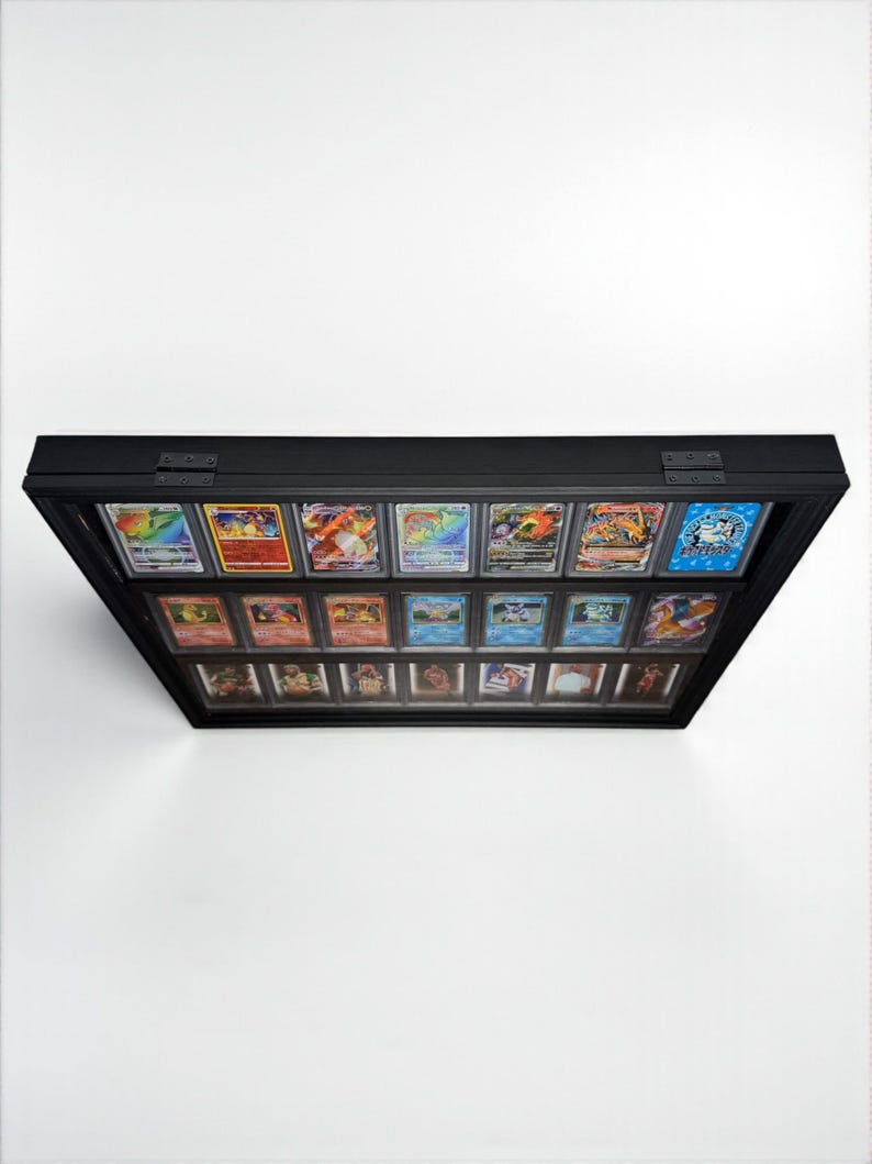 K&ouml;nnte beinhalten: Eine schwarze Vitrine mit Glasfront, die eine Sammlung farbenfroher Sammelkarten pr&auml;sentiert. Die Karten sind in Reihen angeordnet, wobei verschiedene Designs und Illustrationen sichtbar sind. Das Geh&auml;use ist rechteckig und scheint an einer wei&szlig;en Wand montiert zu sein.