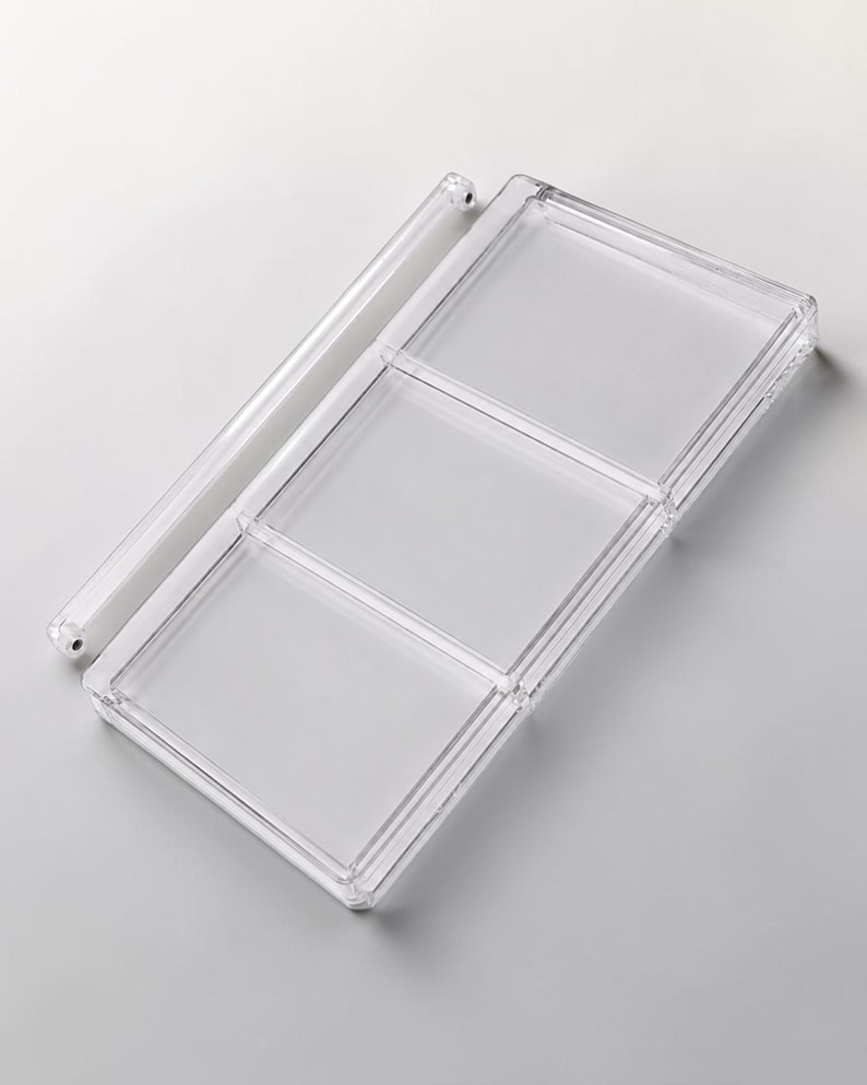 May include: A clear, rectangular plastic organizer with three compartments and a separate, cylindrical bar. The organizer is designed to hold small items and is made of transparent plastic.