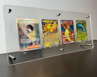 Może przedstawiać: Cztery karty kolekcjonerskie Pokemonów wyświetlane w przezroczystej ramie akrylowej. Karty przedstawiają Zaproszenie Eriki, Zapdosa, Charizarda i Blastoise'a.