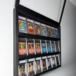 K&ouml;nnte beinhalten: Ein schwarzer Display-Schrank mit drei Regalen, der bewertete Sammelkarten pr&auml;sentiert. Das oberste Regal zeigt farbenfrohe Pok&eacute;mon-Karten, das mittlere Regal zeigt weitere Pok&eacute;mon-Karten und das untere Regal Basketballkarten. Der Schrank ist an einer wei&szlig;en Wand montiert.
