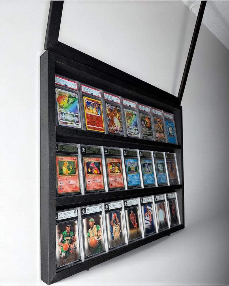 K&ouml;nnte beinhalten: Ein schwarzer Display-Schrank mit drei Regalen, der bewertete Sammelkarten pr&auml;sentiert. Das oberste Regal zeigt farbenfrohe Pok&eacute;mon-Karten, das mittlere Regal zeigt weitere Pok&eacute;mon-Karten und das untere Regal Basketballkarten. Der Schrank ist an einer wei&szlig;en Wand montiert.