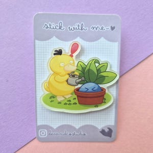 May include: An acrylic pin on a backing card with the text stick with me and deaandcostudio. The pin features a yellow duck-like creature holding a grey watering can, watering a blue round creature inside a brown flower pot with green leaves.