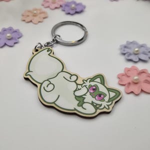 May include: A wooden keychain featuring a cartoon cat design. The cat is white with green accents and pink eyes, lying on its back. The keychain has a silver ring and is surrounded by decorative paper flowers.