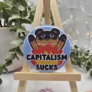 May include: A circular badge with a cartoon character design. The badge has a light blue background with a brown and black character in front of flames. The text "CAPITALISM SUCKS" is printed on the badge.