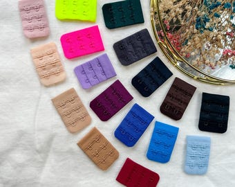 May include: A collection of colorful bra extenders in various shades, including white, pink, beige, green, blue, and purple. Each extender features three sets of hook-and-eye closures. The extenders are arranged on a white surface.