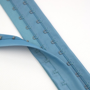May include: A light blue bra extender featuring a long fabric strip with multiple rows of metal hooks and eyes. The fastener is designed for garment adjustment, showcasing a smooth texture and a series of small, silver-toned metal loops.