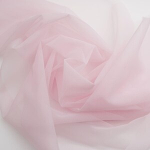 May include: A sheer, pale pink fabric with a fine mesh texture. The material is draped in soft, flowing folds, creating a translucent appearance with varying depths of colour intensity across the layered, lightweight textile surface.