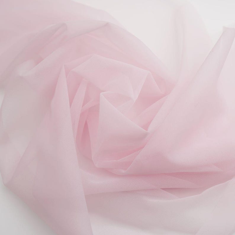 May include: A sheer, pale pink fabric with a fine mesh texture. The material is draped in soft, flowing folds, creating a translucent appearance with varying depths of colour intensity across the layered, lightweight textile surface.