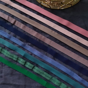 May include: Assortment of ribbon trims in various colors, including pink, brown, blue, green, and teal. The ribbons are arranged in a diagonal pattern on a dark gray surface.