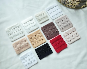 May include: Assortment of 10 bra hook and eye closures in various colours, including white, beige, brown, black, and red. The closures are arranged in a grid pattern on a white surface.