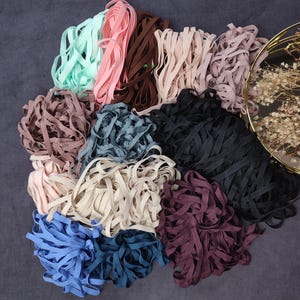 Peut inclure: Assortiment de 12 couleurs de ruban élastique, y compris bleu clair, rose, marron, beige, noir, bleu foncé et violet. Le ruban est disposé en pile sur une surface grise.