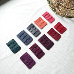 Könnte beinhalten: Sortiment von zehn BH-Trägerverlängerungen in verschiedenen Farben, darunter Burgunderrot, Marineblau, Dunkelgrün und Rot. Jede Verlängerung hat drei Reihen Haken und Ösen.