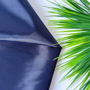 May include: A piece of dark navy blue fabric with a smooth, slightly reflective surface and a fine, tight weave texture. The material is folded to create a sharp diagonal crease, showcasing the deep blue colour and consistent finish.