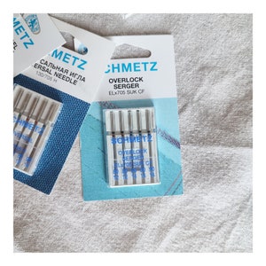 Puede incluir: Un paquete de 6 agujas Schmetz ELx705 SUK CF para remalladoras. Las agujas son plateadas y vienen en un estuche de pl&aacute;stico transparente con una etiqueta azul y blanca.