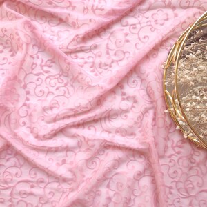 Op de afbeelding: Een close-up van een roze, transparante stof met een bloemmotief. De stof is gedrapeerd, waardoor zachte plooien en schaduwen ontstaan. Een goudkleurige, ronde decoratieve schaal met gedroogde bloemen bevindt zich in de rechterbovenhoek.