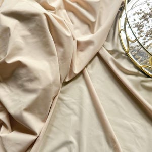 Könnte beinhalten: Nahaufnahme eines beigefarbenen Stoffes mit sanften Falten und Schatten. Der Stoff hat eine glatte Textur und einen subtilen Glanz, der ein Gefühl von Luxus erzeugt. Ein goldgerahmtes Dekotablett mit Trockenblumen im Hintergrund.