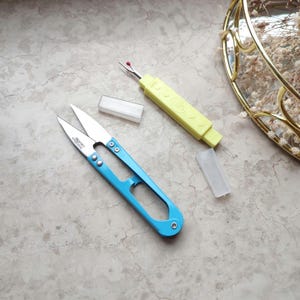 May include: A blue pair of small scissors with a silver blade and a yellow thread cutter with a clear plastic case.