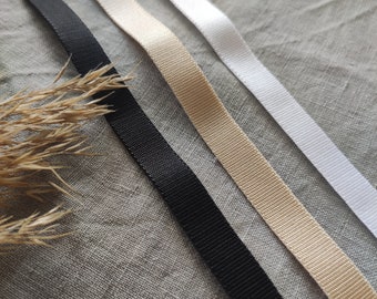 Può includere: Quattro nastri di diversi colori, nero, beige, bianco e crema, sono disposti su una superficie di tessuto strutturato.