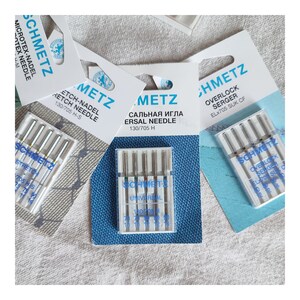 Puede incluir: Primer plano de tres paquetes de agujas para m&aacute;quina de coser. Los paquetes son blancos con detalles azules y llevan el nombre de la marca "Schmetz". Las agujas est&aacute;n envasadas en pl&aacute;stico transparente y est&aacute;n etiquetadas con diferentes tama&ntilde;os y tipos.