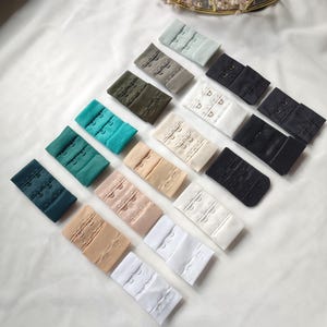 Könnte beinhalten: Sortiment von BH-Trägerverlängerungen in verschiedenen Farben, darunter Weiß, Schwarz, Beige, Grün und Blau. Die Verlängerungen sind aus Stoff und haben drei Haken und Ösen.