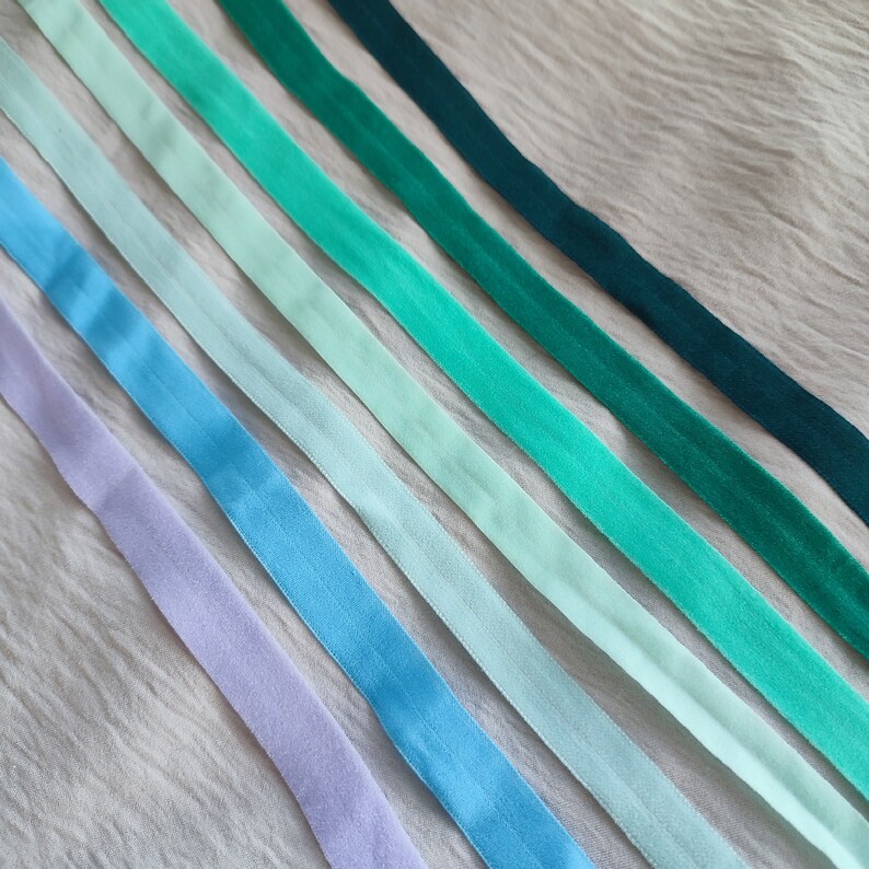 May include: A collection of seven satin ribbons in shades of blue and green, arranged in a gradient pattern on a light beige fabric background.