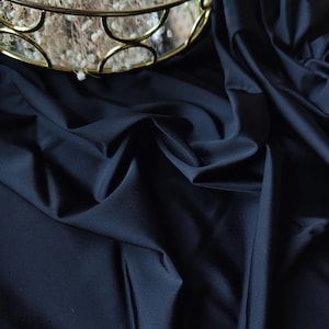 Può includere: Primo piano di tessuto blu navy scuro con morbide pieghe e una leggera lucentezza. Un cestino decorativo in metallo dorato con fiori secchi è visibile in alto a sinistra. La trama del tessuto suggerisce un utilizzo per abbigliamento o arredamento.