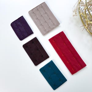 Op de afbeelding: Assortiment van bh-verlengers in diverse kleuren, waaronder paars, taupe, bruin, rood en blauwgroen. Elke verlenger heeft drie sets haak-en-oog sluitingen. Deze stoffen verlengers zijn ontworpen om extra lengte aan bh-banden toe te voegen.