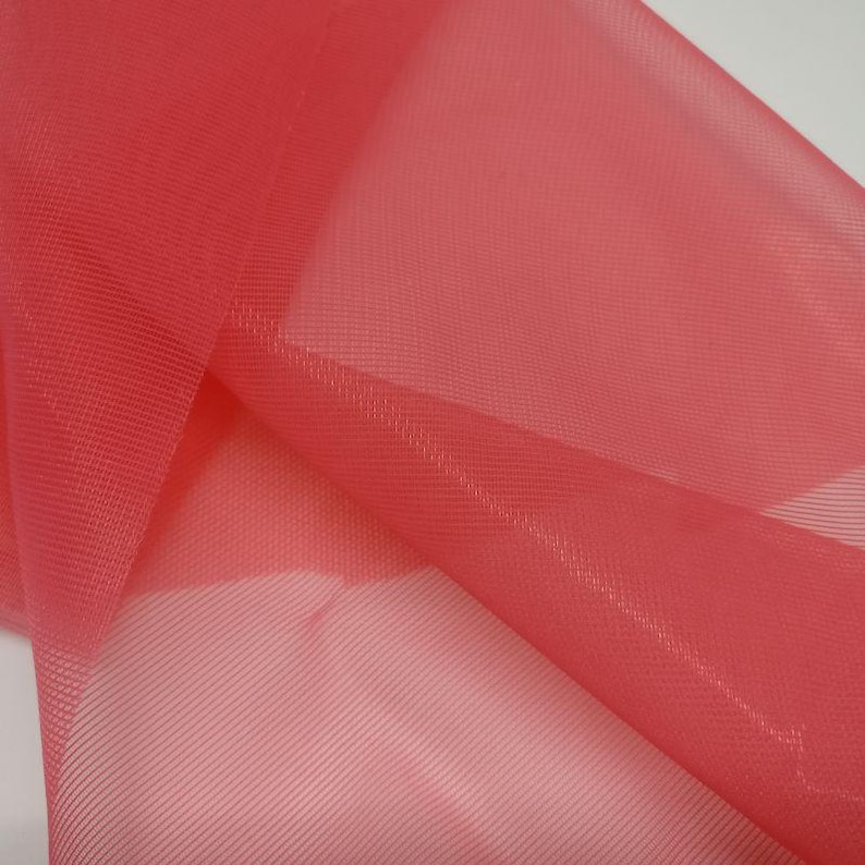 May include: A sheer, coral-pink fabric with a fine, grid-like mesh texture. The material is lightweight and translucent, featuring a consistent woven pattern throughout the surface, creating soft folds and layers of semi-transparent colour.