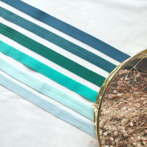 May include: Five different shades of blue and green elastic ribbon arranged in a gradient from dark blue to light blue. The ribbon is lying on a white surface.