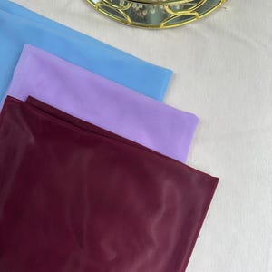 May include: Three rectangular pieces of fabric stacked on top of each other. The top layer is light blue, the middle layer is light purple, and the bottom layer is a deep burgundy red. Each piece of fabric features a fine, vertical ribbed texture.