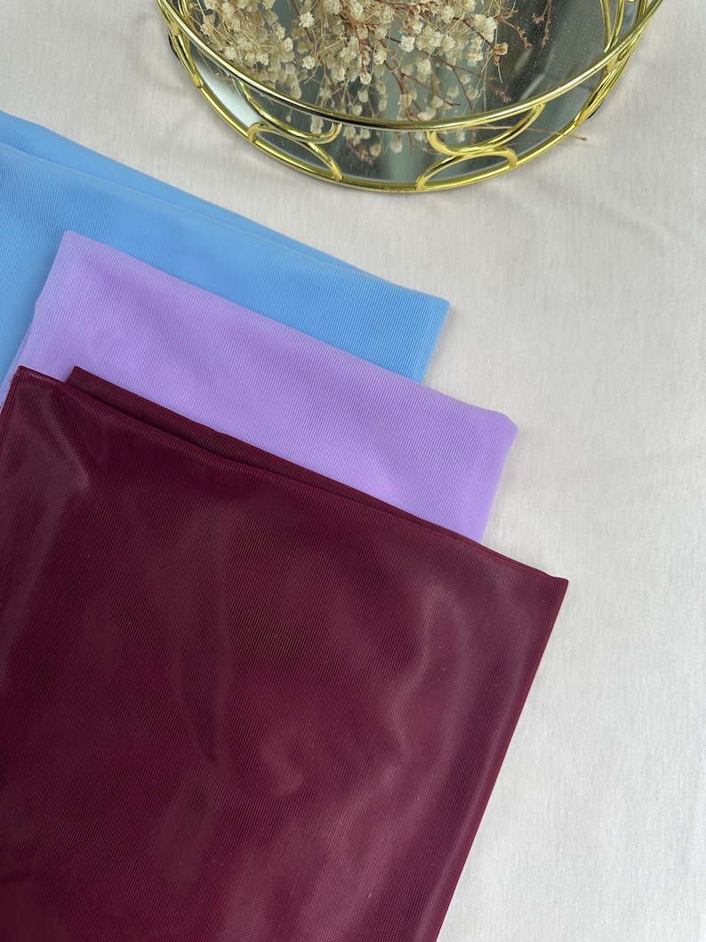 May include: Three rectangular pieces of fabric stacked on top of each other. The top layer is light blue, the middle layer is light purple, and the bottom layer is a deep burgundy red. Each piece of fabric features a fine, vertical ribbed texture.