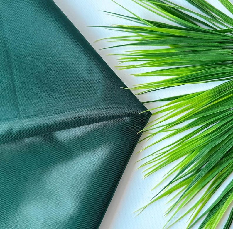 May include: A smooth, dark forest green fabric with a subtle, fine-grained texture. The material is folded to create soft, diagonal lines and shadows across its surface, contrasting with the bright green, pointed artificial grass blades.